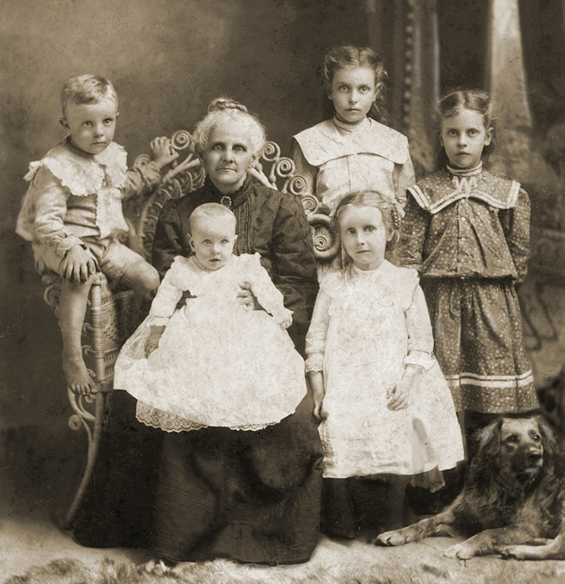 Family portrait in sepia showing an older woman seated with five children and a dog against a studio backdrop