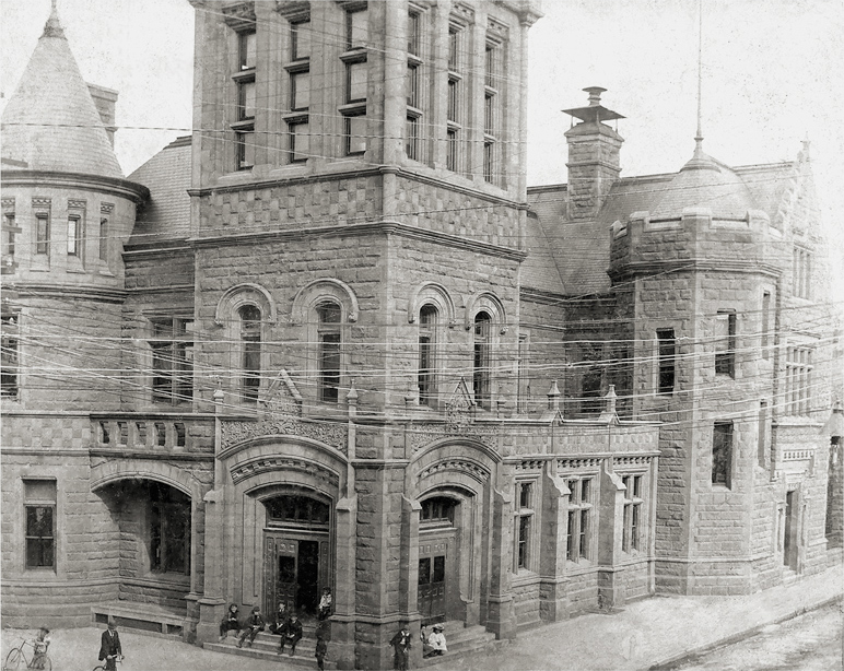 Stone building in black and white, with towers, ornate arched entrances and overhead wires, beside pedestrians on a street