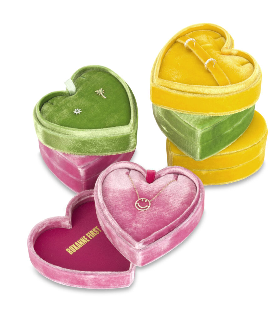Heart-shaped velvet jewellery boxes in pink, green and yellow displaying earrings and a smiley pendant necklace on a white background